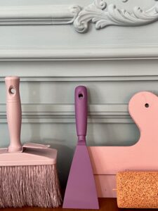 Professionelle Malerwerkzeuge (Spachtel, Pinsel, Rolle) vor einer Wand mit filigranen Stuckarbeiten und Wandpaneelen. Zeigt Malerarbeiten Wien.